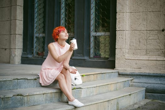 drinks coffee from a cup. woman with dyed red hair in a pale pink dress drinking coffee, holding a paper cup of coffee to go, sitting on the steps outside a summer day.