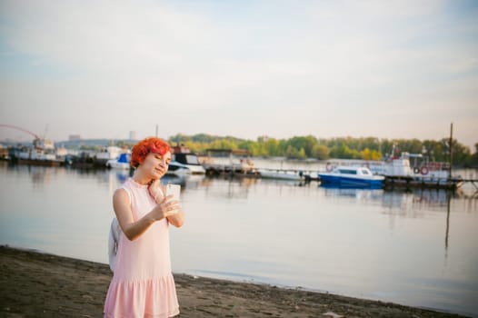 girl in pale pink dress with red hair and backpack walking along river bank, pictures of themselves on their mobile camera phone, against backdrop of boats moored on a warm summer day