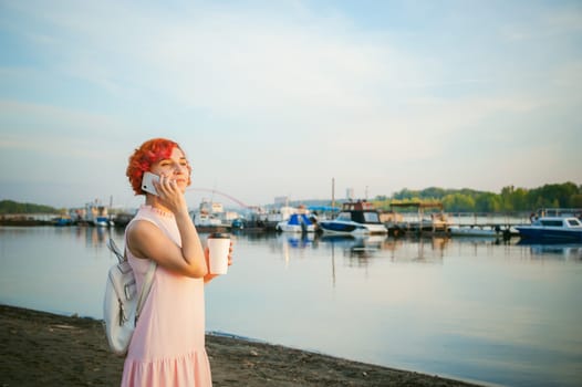 girl in pale pink dress with red hair and backpack walking along river bank, talking on the phone and drinking coffee from a cardboard cup, against backdrop of boats moored on a warm summer day