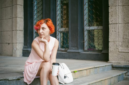 drinks coffee from a cup. woman with dyed red hair in a pale pink dress drinking coffee, holding a paper cup of coffee to go, sitting on the steps outside a summer day.