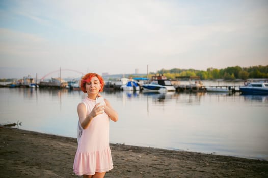 girl in pale pink dress with red hair and backpack walking along river bank, pictures of themselves on their mobile camera phone, against backdrop of boats moored on a warm summer day