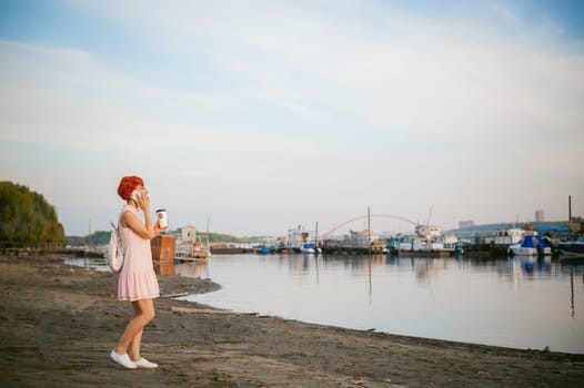 girl in pale pink dress with red hair and backpack walking along river bank, talking on the phone and drinking coffee from a cardboard cup, against backdrop of boats moored on a warm summer day