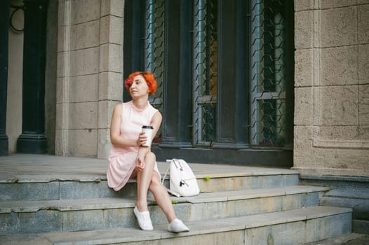 drinks coffee from a cup. woman with dyed red hair in a pale pink dress drinking coffee, holding a paper cup of coffee to go, sitting on the steps outside a summer day.