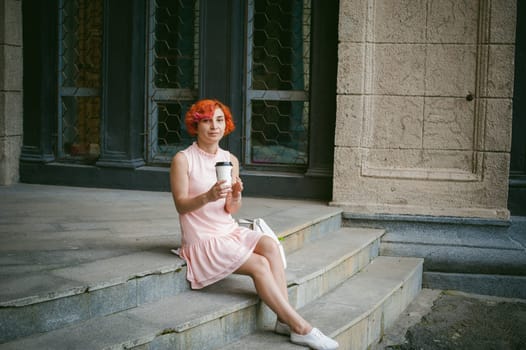 drinks coffee from a cup. woman with dyed red hair in a pale pink dress drinking coffee, holding a paper cup of coffee to go, sitting on the steps outside a summer day.