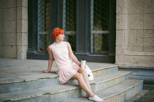 drinks coffee from a cup. woman with dyed red hair in a pale pink dress drinking coffee, holding a paper cup of coffee to go, sitting on the steps outside a summer day.