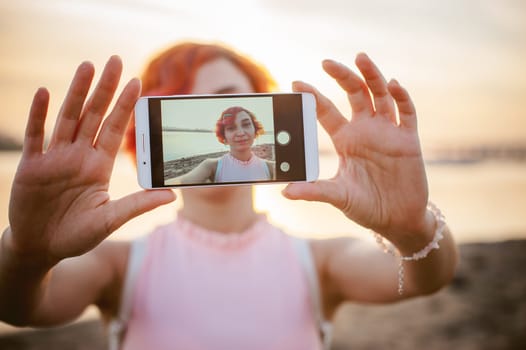 girl in pale pink dress with red hair and backpack walking along river bank, pictures of themselves on their mobile camera phone, against backdrop of boats moored on a warm summer day