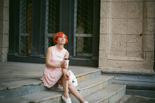 drinks coffee from a cup. woman with dyed red hair in a pale pink dress drinking coffee, holding a paper cup of coffee to go, sitting on the steps outside a summer day.