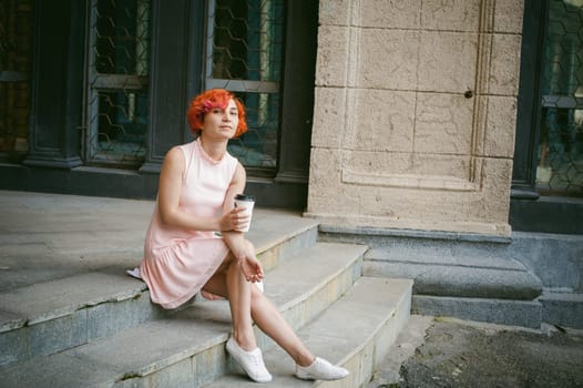 drinks coffee from a cup. woman with dyed red hair in a pale pink dress drinking coffee, holding a paper cup of coffee to go, sitting on the steps outside a summer day.