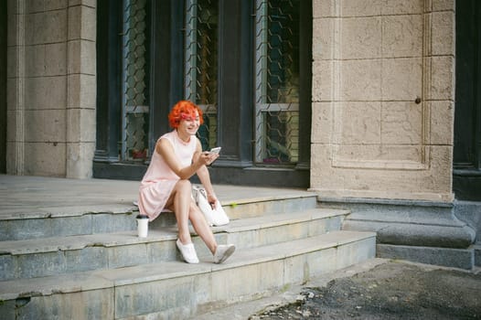 girl doing selfie. woman with dyed red hair in a pale pink dress with a backpack white color drinking coffee, pictures of themselves on their mobile camera phone, sitting on the stairs summer day