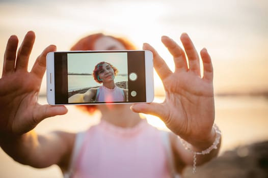 girl in pale pink dress with red hair and backpack walking along river bank, pictures of themselves on their mobile camera phone, against backdrop of boats moored on a warm summer day