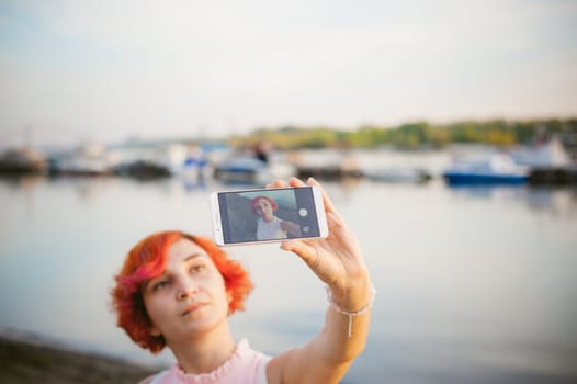 girl in pale pink dress with red hair and backpack walking along river bank, pictures of themselves on their mobile camera phone, against backdrop of boats moored on a warm summer day