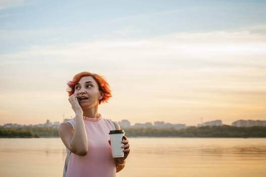 girl in pale pink dress with red hair and backpack walking along river bank, talking on the phone and drinking coffee from a cardboard cup, against backdrop of boats moored on a warm summer day