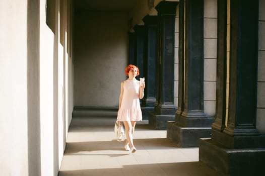 girl in a pale pink dress with dyed red hair, a white backpack in hand, drinks coffee from a cup, walking between light and shadow on the columns sunny summer day