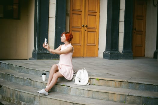 girl doing selfie. woman with dyed red hair in a pale pink dress with a backpack white color drinking coffee, pictures of themselves on their mobile camera phone, sitting on the stairs summer day