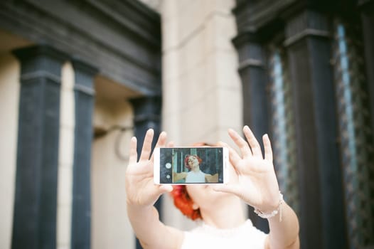 portrait of a girl doing selfie. girl in light pink dress with red dyed hair, pictures of themselves on their mobile camera phone, sitting on the stairs summer day