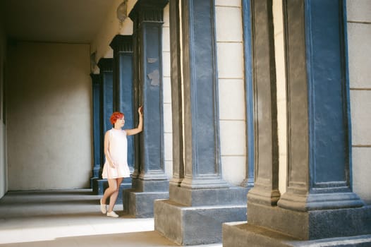 girl in a pale pink dress with dyed red hair, a white backpack in hand, walking between light and shadow on the columns sunny summer day