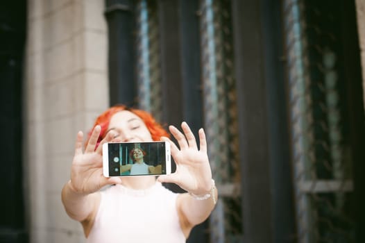 portrait of a girl doing selfie. girl in light pink dress with red dyed hair, pictures of themselves on their mobile camera phone, sitting on the stairs summer day