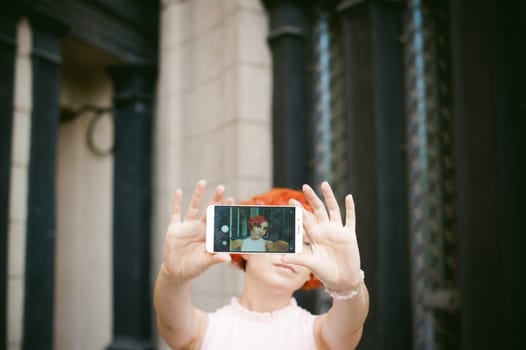 portrait of a girl doing selfie. girl in light pink dress with red dyed hair, pictures of themselves on their mobile camera phone, sitting on the stairs summer day