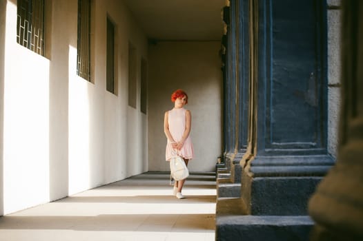 girl in a pale pink dress with dyed red hair, a white backpack in hand, walking between light and shadow on the columns sunny summer day