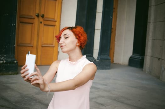 portrait of a girl doing selfie. girl in light pink dress with red dyed hair, pictures of themselves on their mobile camera phone, sitting on the stairs summer day