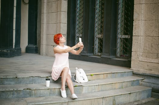girl doing selfie. woman with dyed red hair in a pale pink dress with a backpack white color drinking coffee, pictures of themselves on their mobile camera phone, sitting on the stairs summer day