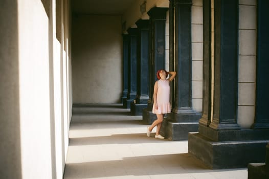girl in a pale pink dress with dyed red hair, a white backpack in hand, walking between light and shadow on the columns sunny summer day