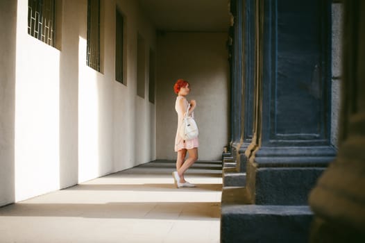 girl in a pale pink dress with dyed red hair, a white backpack in hand, walking between light and shadow on the columns sunny summer day