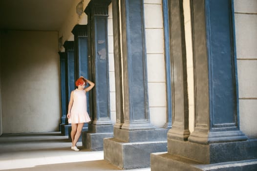 girl in a pale pink dress with dyed red hair, a white backpack in hand, walking between light and shadow on the columns sunny summer day
