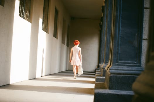 girl in a pale pink dress with dyed red hair, a white backpack in hand, walking between light and shadow on the columns sunny summer day
