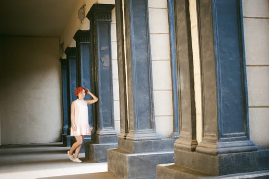 girl in a pale pink dress with dyed red hair, a white backpack in hand, walking between light and shadow on the columns sunny summer day