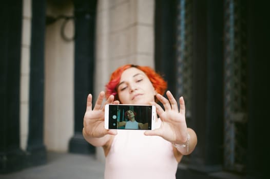 portrait of a girl doing selfie. girl in light pink dress with red dyed hair, pictures of themselves on their mobile camera phone, sitting on the stairs summer day