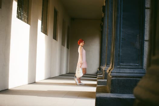 girl in a pale pink dress with dyed red hair, a white backpack in hand, walking between light and shadow on the columns sunny summer day