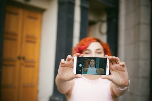 portrait of a girl doing selfie. girl in light pink dress with red dyed hair, pictures of themselves on their mobile camera phone, sitting on the stairs summer day