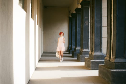 girl in a pale pink dress with dyed red hair, a white backpack in hand, drinks coffee from a cup, walking between light and shadow on the columns sunny summer day