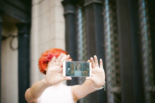 portrait of a girl doing selfie. girl in light pink dress with red dyed hair, pictures of themselves on their mobile camera phone, sitting on the stairs summer day