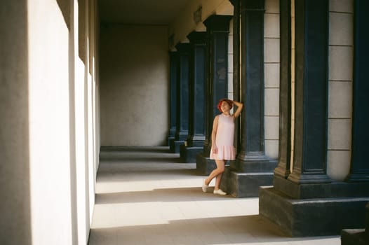 girl in a pale pink dress with dyed red hair, a white backpack in hand, walking between light and shadow on the columns sunny summer day