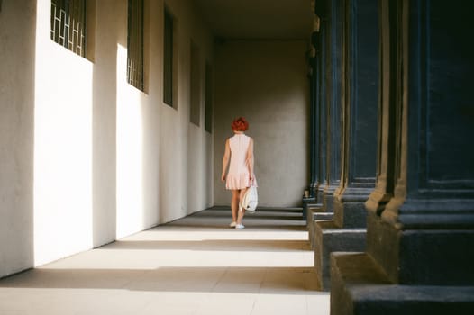 girl in a pale pink dress with dyed red hair, a white backpack in hand, walking between light and shadow on the columns sunny summer day