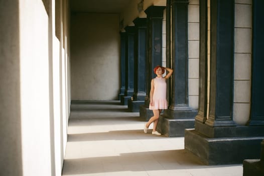 girl in a pale pink dress with dyed red hair, a white backpack in hand, walking between light and shadow on the columns sunny summer day