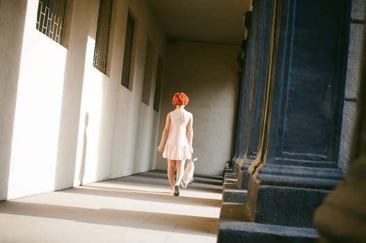 girl in a pale pink dress with dyed red hair, a white backpack in hand, walking between light and shadow on the columns sunny summer day