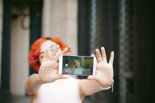 portrait of a girl doing selfie. girl in light pink dress with red dyed hair, pictures of themselves on their mobile camera phone, sitting on the stairs summer day