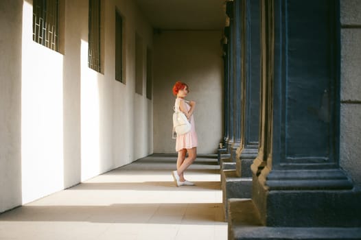 girl in a pale pink dress with dyed red hair, a white backpack in hand, walking between light and shadow on the columns sunny summer day