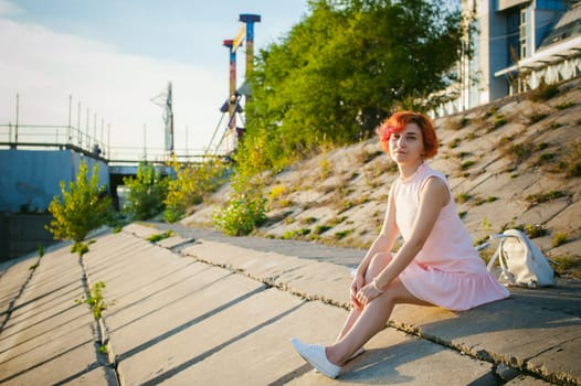girl in pale pink dress with red hair and backpack walking along river bank, sitting on the sandy shore