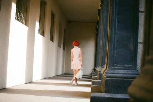 girl in a pale pink dress with dyed red hair, a white backpack in hand, walking between light and shadow on the columns sunny summer day