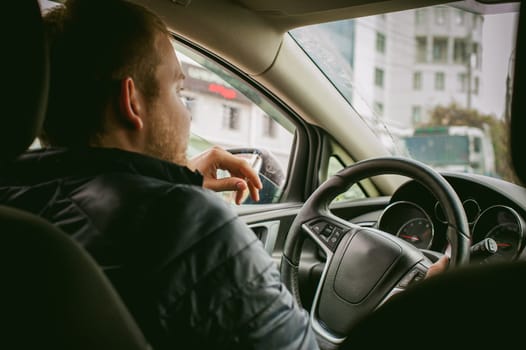 man drives a car. A guy in a jacket sits behind the wheel of a car traveling on road in traffic jam, smoking a cigarette. Photo taken off the seat, due to the driver's back