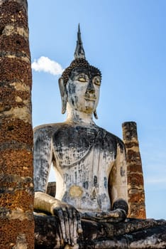 Ancient Buddha statue sitting among the ruins under the bright sky of Wat Maha That temple in Sukhothai Historical Park is an old city and famous tourist attraction of Sukhothai Province, Thailand