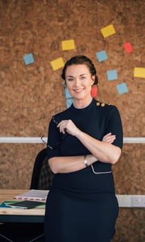 Portrait of a beautiful woman in her workplace. She is standing in front of a corkboard and is smartly dressed, smiling for the camera.