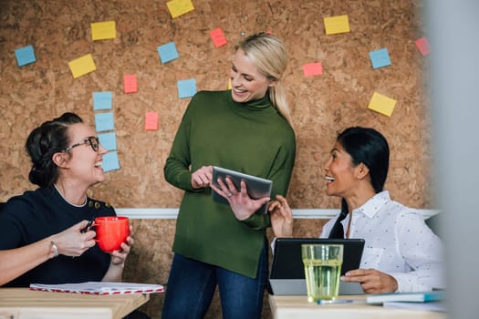 Women are working together in an office. They are talking and laughing as they use digital tablets.