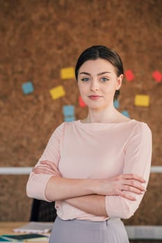 Portrait of a female office worker. She is standing in front of a cork board, smartly dressed with her arms folded and is smiling for the camera.