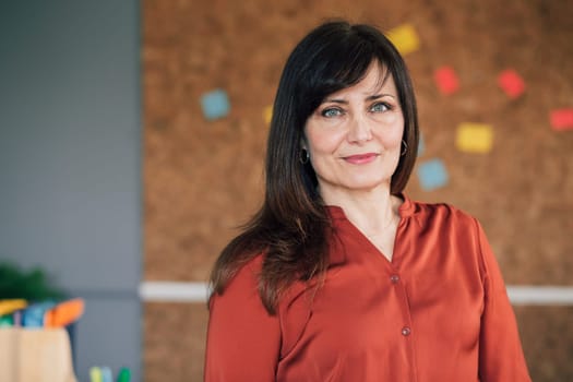 Portrait of a mature, female business owner in her office. She is standing in front of a cork board and is smartly dressed, smiling for the camera.