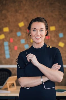 Portrait of a beautiful woman in her workplace. She is standing in front of a corkboard and is smartly dressed, smiling for the camera.
